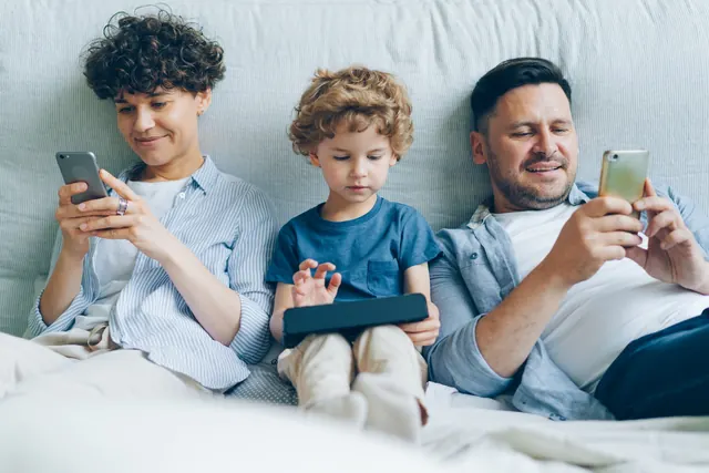 Táta se dvěma syny na gauči, každý s mobilem nebo tabletem