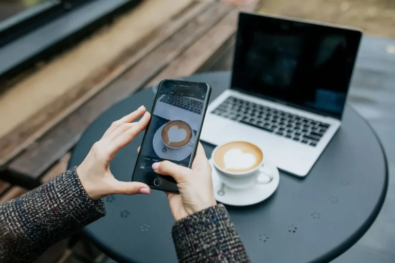 Ruce drží iPhone u stolku s kávou a notebookem